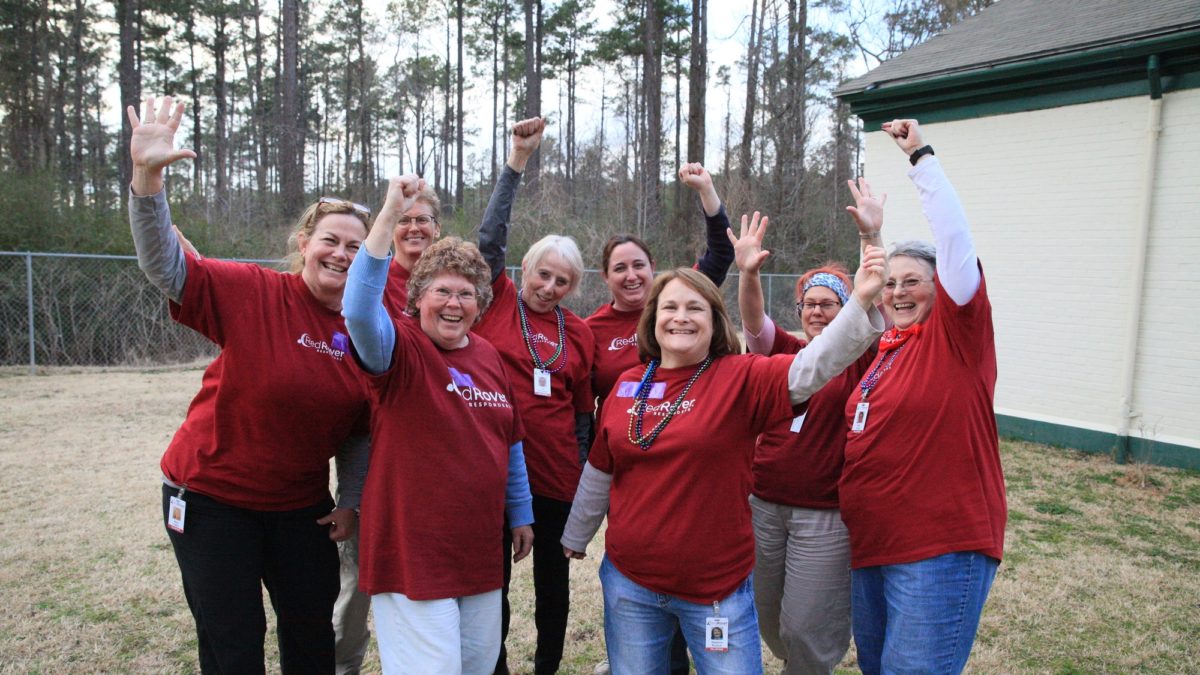 Group of eight women in RedRover Responders t-shirts outside, smiling, waving their hands triumphantly above their heads