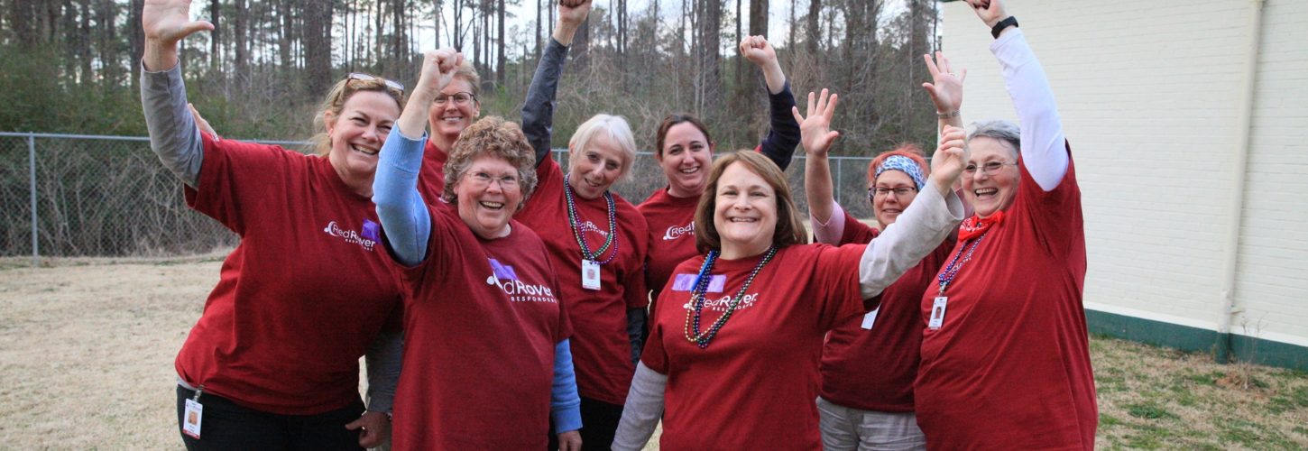Group of eight women in RedRover Responders t-shirts outside, smiling, waving their hands triumphantly above their heads