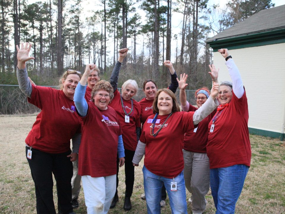 Group of eight women in RedRover Responders t-shirts outside, smiling, waving their hands triumphantly above their heads