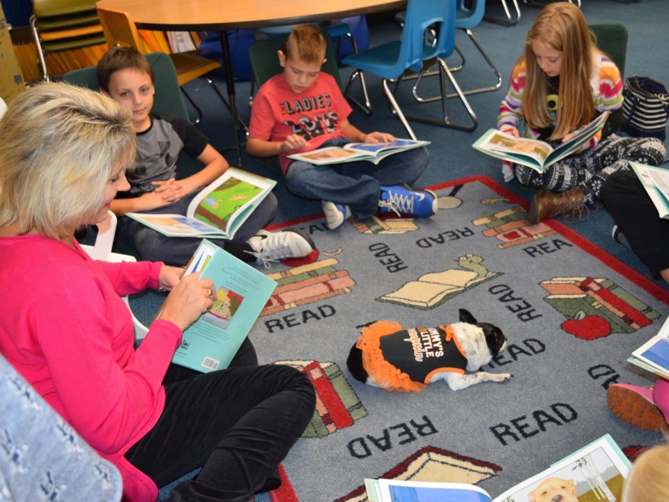 Teacher sitting on rug with several students. Each person is holding a copy of the book Buddy Unchained, a book about a dog, and smiling.
