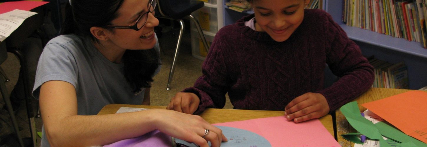Teacher with glasses smiling and kneeling next to a student at a desk. The student is working on an animal-themed drawing and smiling.
