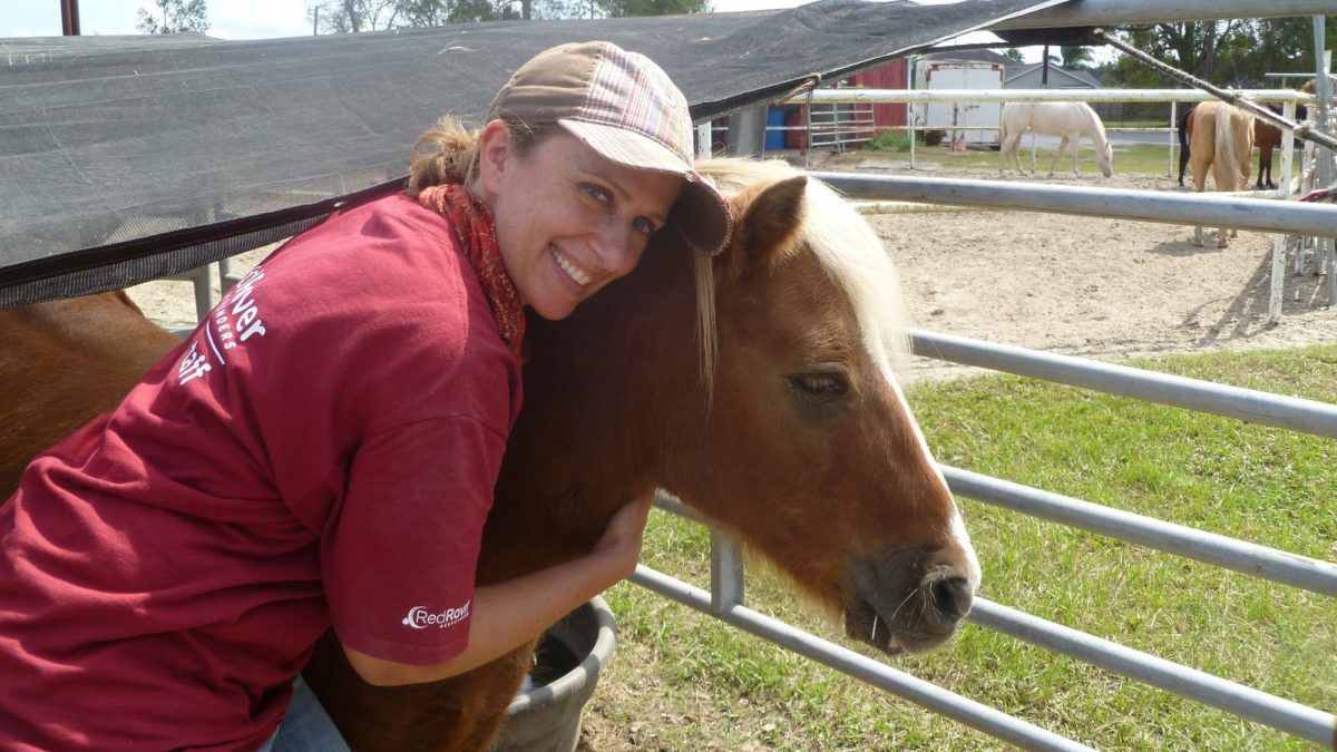 A woman in a RedRover Responders tshirt wearing a tan hat hugs a brown pony