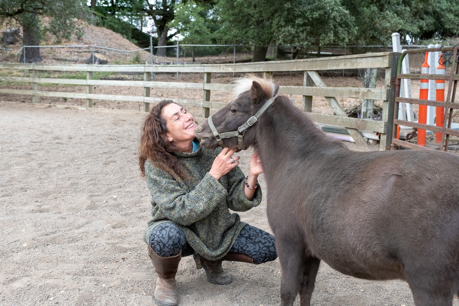 A woman with long, wavy brown hair in a green sweater, blue tights, and brown boots squats down to pet the face of a small brown horse in a paddock