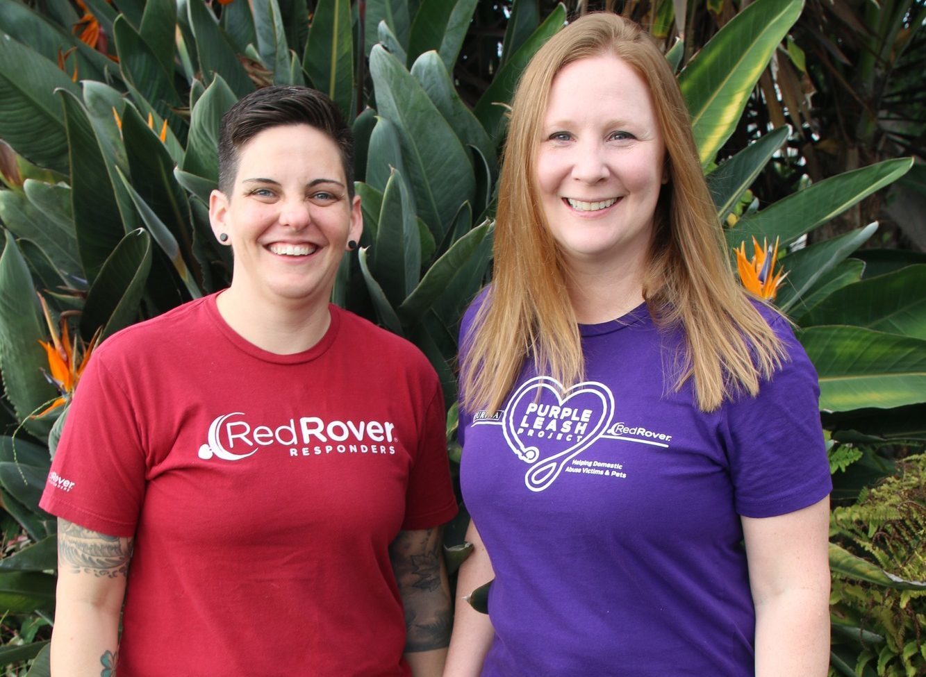 A smiling brunette woman with a cropped haircut wearing a red t-shirt that has the RedRover logo on the chest stands besides a smiling blonde woman wearing a purple tshirt with the Purple Leash Project logo on the chest.