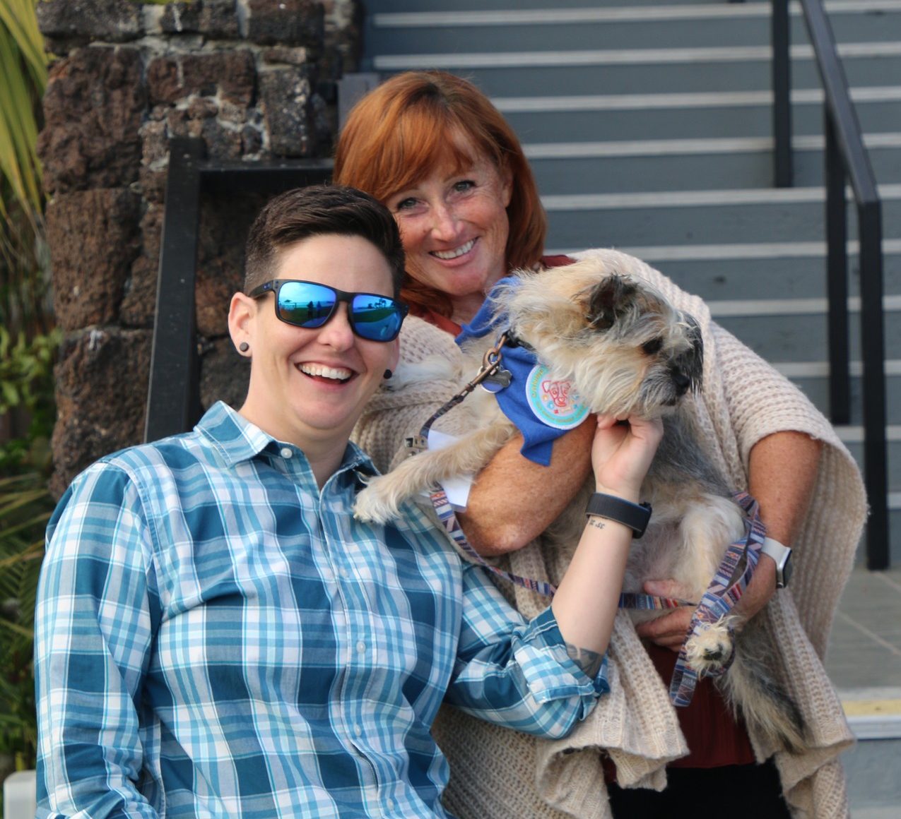 A brunette woman with a cropped haircut, sunglasses, and a blue plaid shirt stands beside a red-headed woman in a cream-colored sweater holding a small, scruffy, cream-colored dog. They are standing in front of an outdoor staircase and smiling.