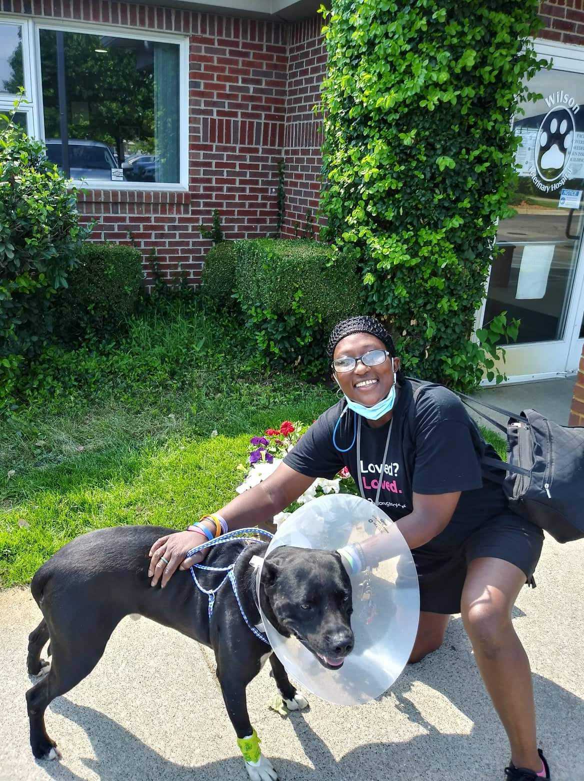 A black dog with a cone stands beside her smiling owner