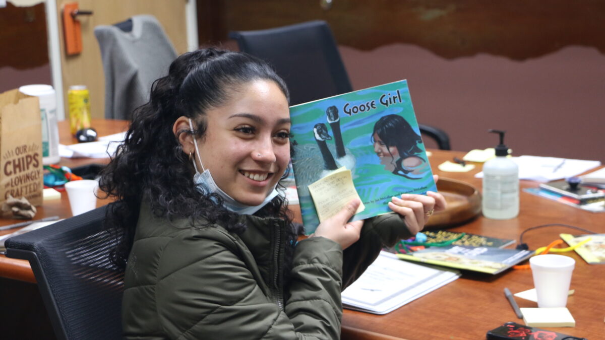 A Native teen smiles as they holds up a children's book titled Goose Girl. They are wearing a dark green jacket and have their black hair pulled back into a ponytail.