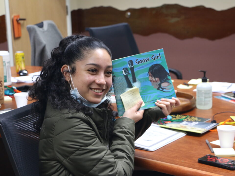 A Native teen smiles as they holds up a children's book titled Goose Girl. They are wearing a dark green jacket and have their black hair pulled back into a ponytail.
