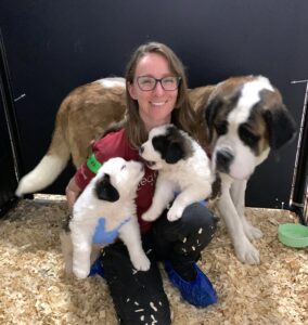 woman with glasses and red shirt holding two puppies next to their mother