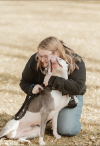 Woman wearing glasses and a black longsleeve shirt hugging her bully dog in a field