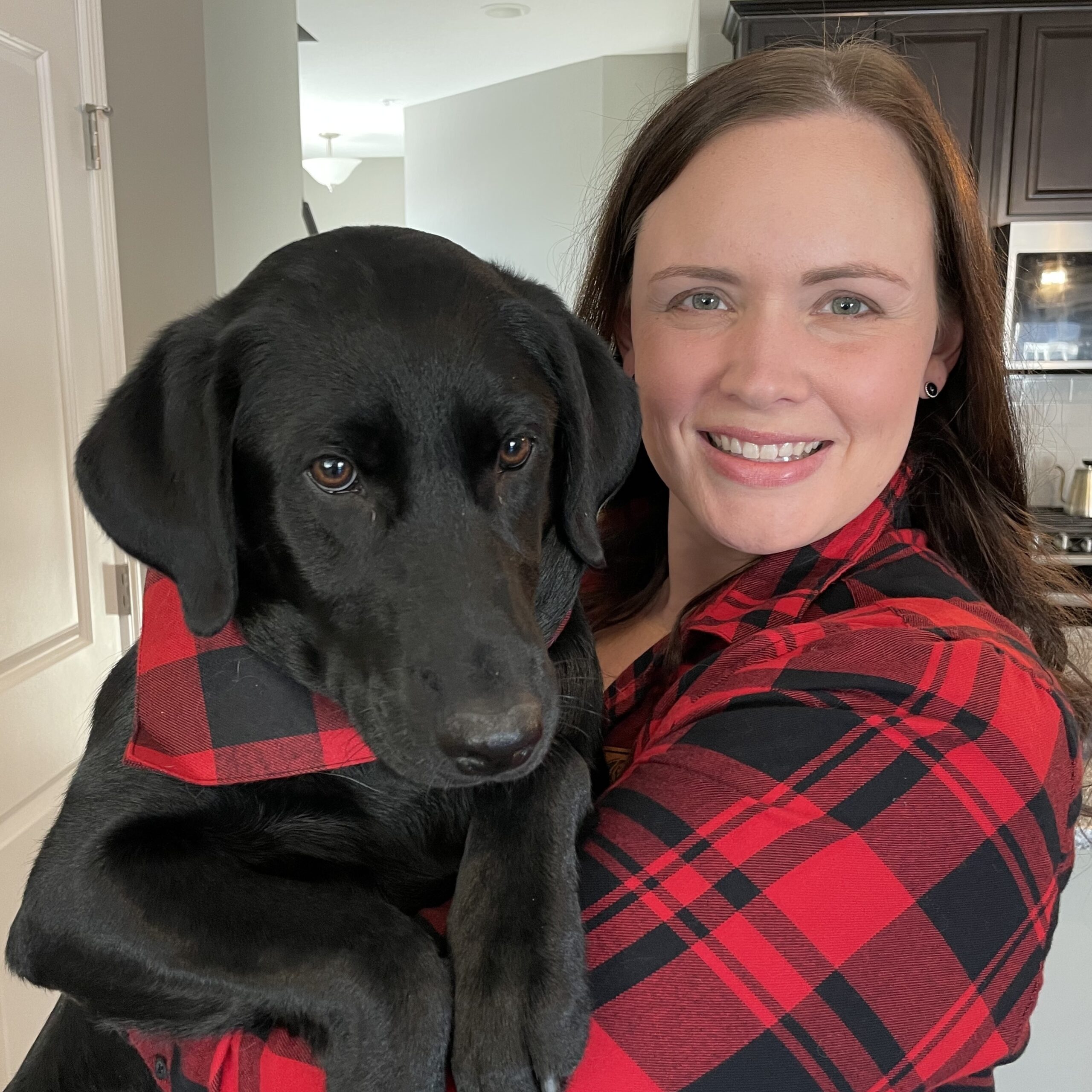 Woman with brown hair and black and red flannel shirt holds her dog, a black labrador